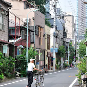 Man walks a bicycle down a narrow residential street with high-rise buildings in the background.